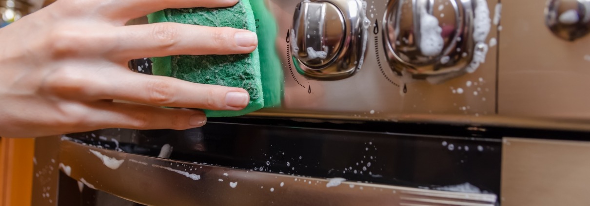 feminine hand with a green sponge washes the knobs of a stainless steel stove.