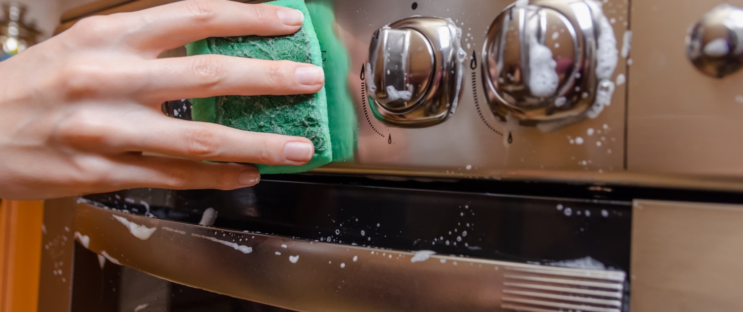 feminine hand with a green sponge washes the knobs of a stainless steel stove.