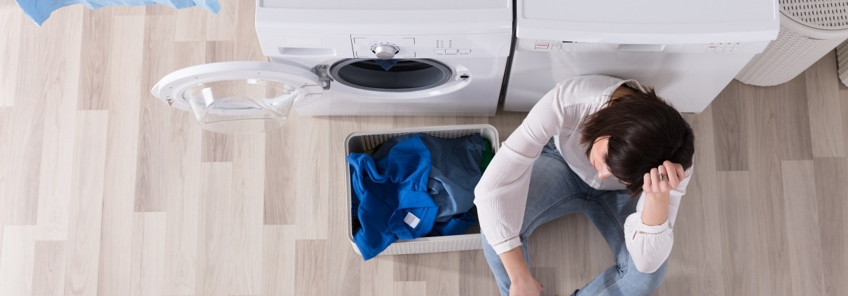 Upset woman sitting in front of washer and dryer beside basket of dirty laundry