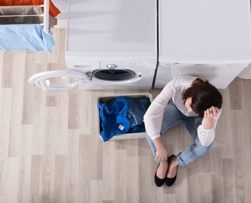 Upset woman sitting in front of washer and dryer beside basket of dirty laundry