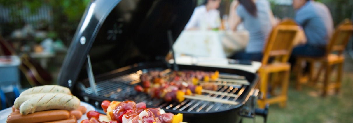 Close up of skewers on a grill with people in the background enjoying the summer