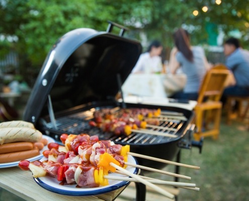 Close up of skewers on a grill with people in the background enjoying the summer