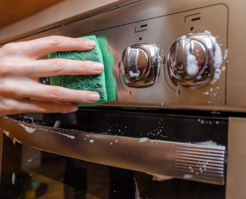 feminine hand with a green sponge washes the knobs of a stainless steel stove.